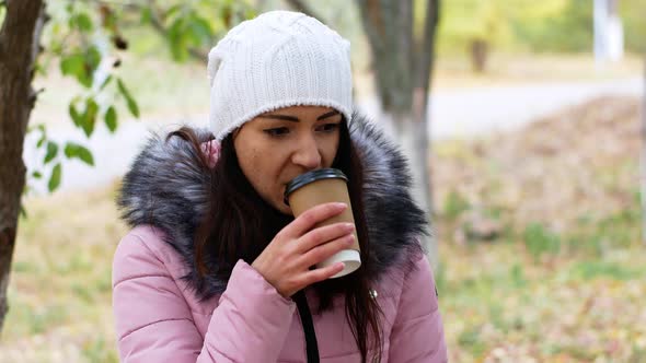 A beautiful woman with long dark hair sits on a park bench and drinks delicious aromatic coffee