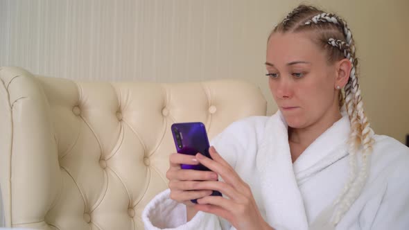 Young woman in white bathrobe uses mobile phone while sitting on armchair.