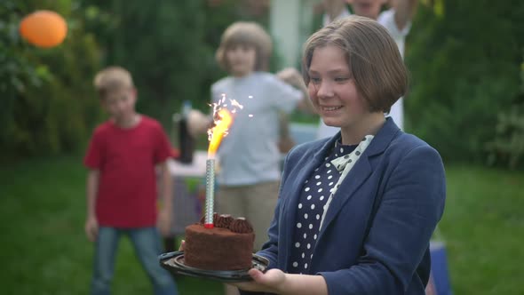 Excited Caucasian Teenage Girl Admiring Birthday Candle Burning in Cake with Group of Blurred