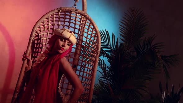 a Blonde in a Turban with a Red Scarf Sits in a Wicker Hanging Chair and Moves Hands Beautifully in