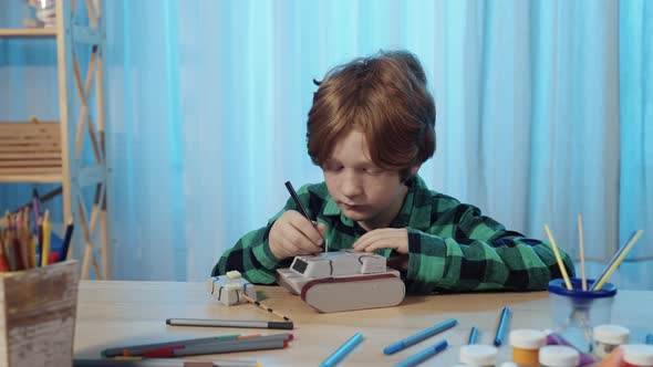 Little Schoolboy Sitting at Table in Room and Paints Toy Tank