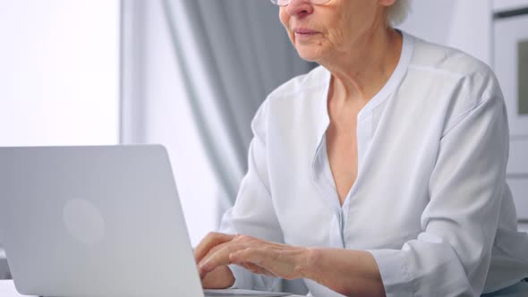 Old woman manager in glasses types on grey laptop keyboard sitting at large white table