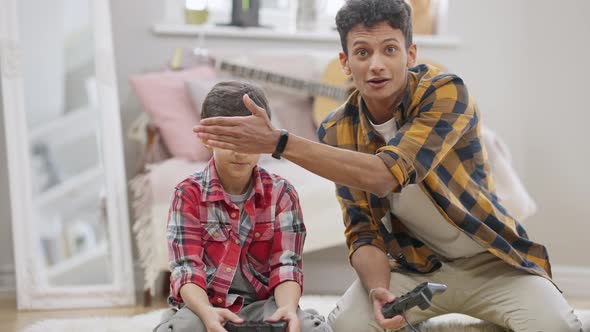 Laughing Middle Eastern Teenager Closing Eyes of Little Brother Playing Video Game Using Game