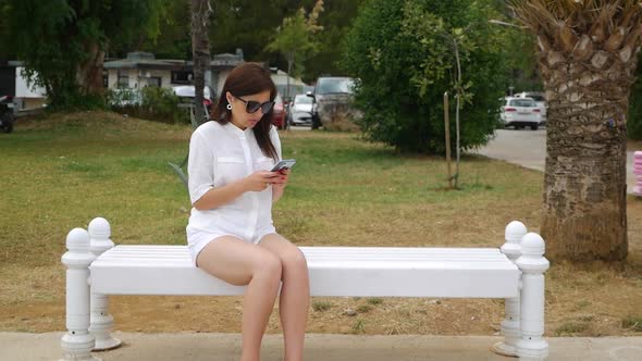 The Girl is Sitting on a Bench and Uses the Phone