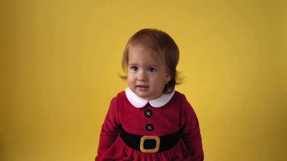 Cute Happy Cheerful Chubby Toddler Baby Girl in Santa Suit Looking On Camera At Yellow Background