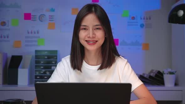 Happy excited Asian business woman making thumb up sign and working on laptop on the table at night.