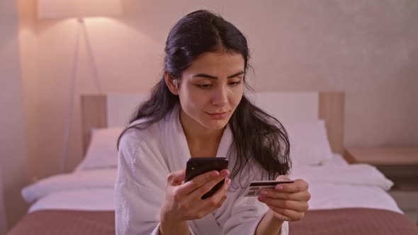 Smiling Young Woman Customer Holding Credit Card and Smartphone Sitting on Couch at Home