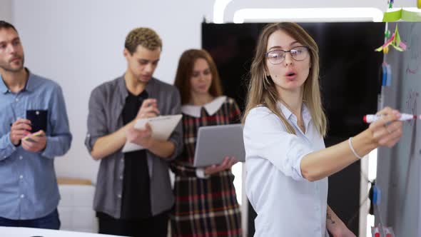 Young Woman Ofice Worker Standing and Writing on White Board an Talking with Her Colleagues