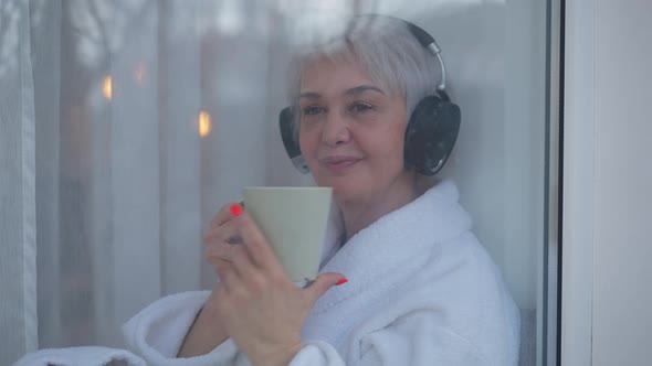 Shooting Thorough Window Glass Happy Mature Woman in Headphones Enjoying Taste of Morning Coffee at