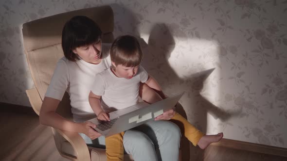 Mother and Her Toddler Boy Sit Together on Chair with Laptop. Woman Tries To Remote Work, but Kid Is