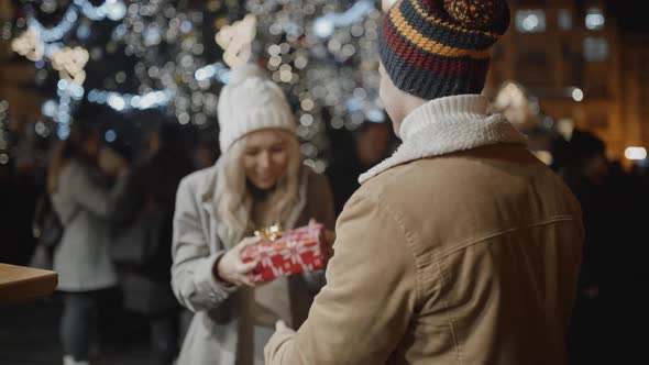 Man Standing Back to Camera Presents a Gift to Her Girlfriend at Xmas Night at Old Town Square