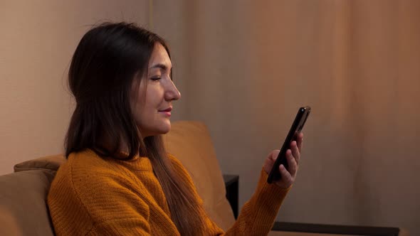 Brunette in Pullover Sits on Sofa and Types on Smartphone