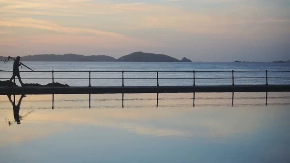 A Man Carrying A Camera Stand Walking Beside The Swimming Pool Overlooking The Island And The Waters