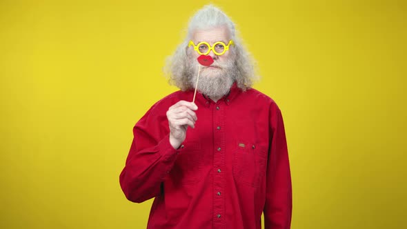 Joyful Senior Retiree in Yellow Eyeglasses Posing with Toy Lips and Waving at Camera