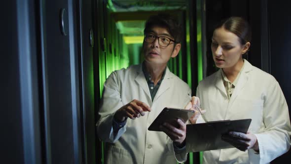 Diverse female and male it technicians in lab coats using tablet and laptop checking computer server