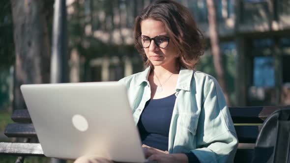 A Young Woman Student Sitting on a Bench in the Park and Typing on Her Laptop