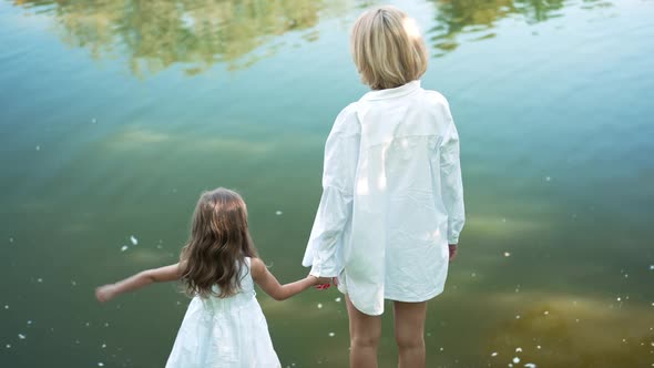 Happy Mother and Daughter Holding Hands Standing at Background of Lake Waters in Summer Spring Park