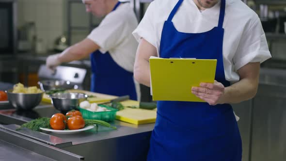 Unrecognizable Chef in Apron Writing New Recipe Standing in Kitchen with Blurred Cook Working at