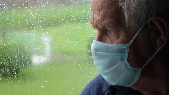 Sad Old Man In Protective Medical Mask Looks Out Window At Street When It Rains