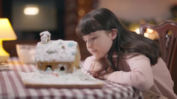 Side View Happy Cute Girl Admiring Cooked Decorated Gingerbread House Sitting in Kitchen at Home