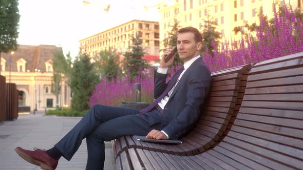 Young Busy Businessman Sits on Bench with Purple Flowers in Flower Beds Behind Him and Use Mobile