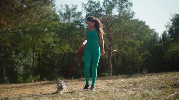 Beautiful athletic woman in sporty turquoise overalls walks with her fluffy cat on a leash in forest