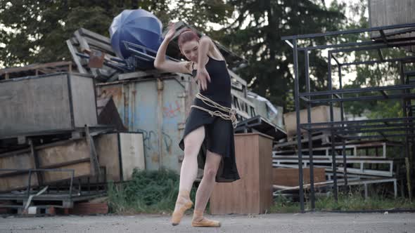 Wide Shot of Slim Elegant Ballerina in Black Dress Bound with Rope Dancing on Urban Slum