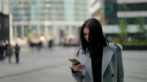 a Brunette in a Gray Coat is Typing on Phone and Walking Against the Background of Blurry Modern