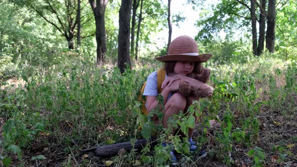 Little Girl with a Teddy Bear and Yellow Backpack Sits Alone in the Thicket