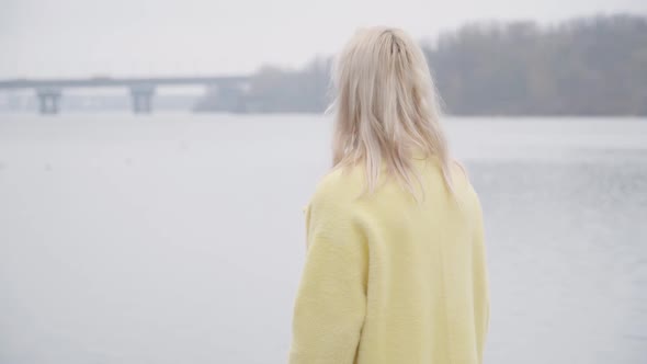 Lonely Blond Caucasian Woman in Yellow Coat Standing on Riverbank and Looking Away