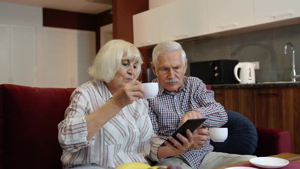 Senior Pensioner Couple with Digital Tablet Pc Computer at Home. Resting on Sofa in Cozy Living Room