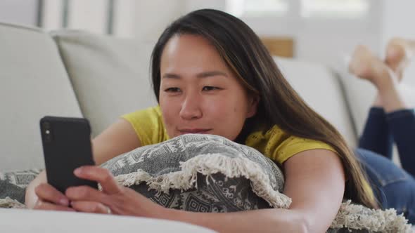 Happy asian woman lying on sofa, resting with smartphone at home