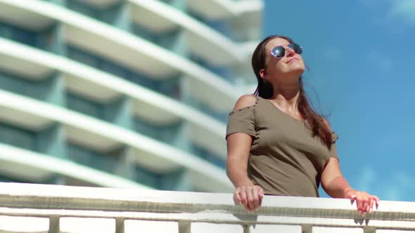 Attractive Smiling Tanned Woman Wearing Sunglasses Relaxing on Balcony of Luxury Modern Hotel