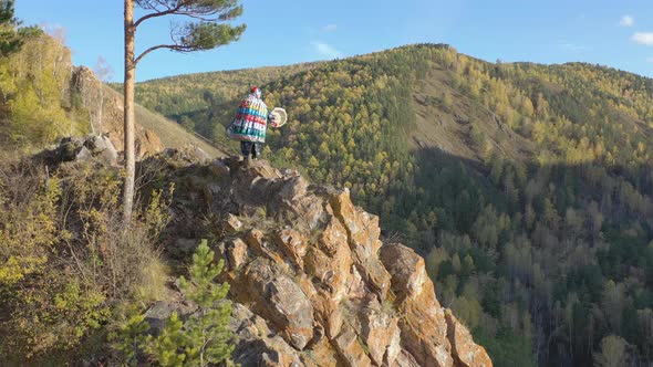 Aerial Shot of an Old Wizard in a Bright Shaman Costume Dancing with a Tambourine on a Mountain Top