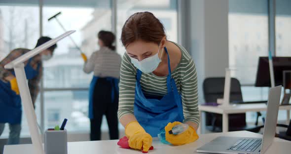 Portrait of Young Female Worker in Uniform and Safety Mask Cleaning Desk with Rag and Spray in