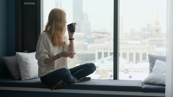 woman drinking tea while sitting on the windowsill overlooking the city