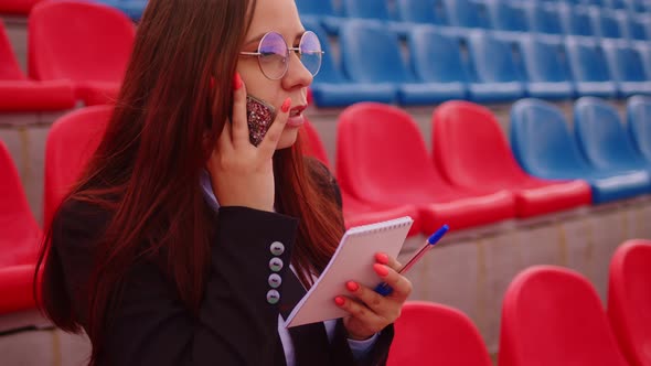 Young Woman in Glasses with Notepad Pen Talking on Mobile Phone Sitting on Stadium Bleachers