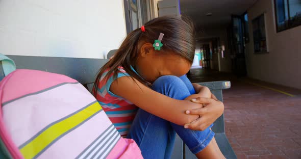 Side view of African American schoolgirl with head down sitting alone on bench in school corridor 4k