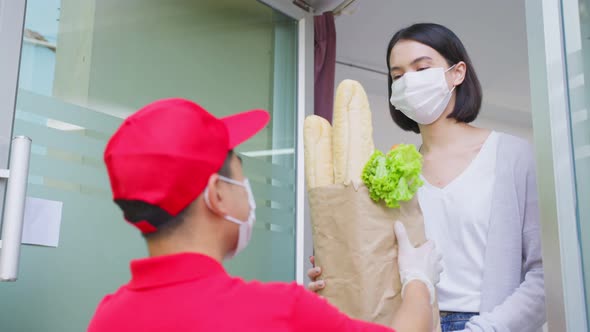 Asian deliver man wearing face mask in red uniform handling bag of food, groceries to young woman.