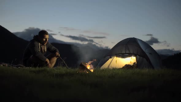 Young Man Warming with Camp Fire in Nature Mountain Outdoor Camping Scene at Night