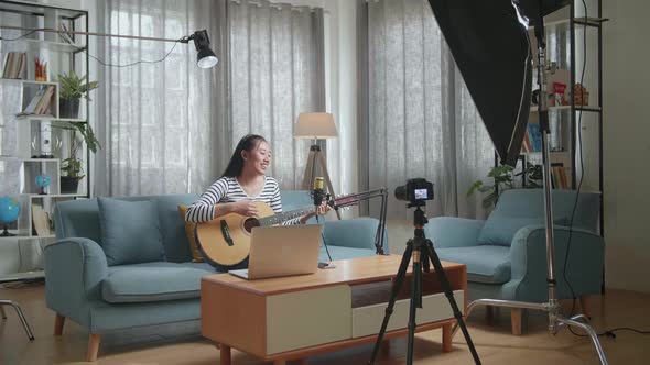 Asian Woman With A Laptop Singing And Playing A Guitar While Live Stream At Home