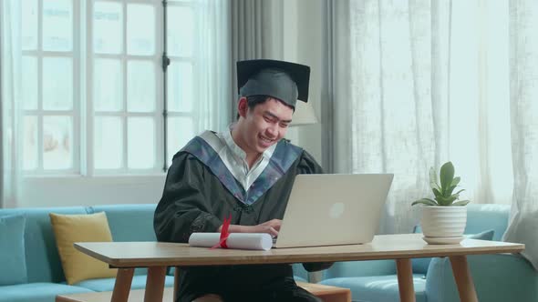 Happy Asian Man Wearing A Graduation Gown And Cap Use Laptop Computer In Living Room