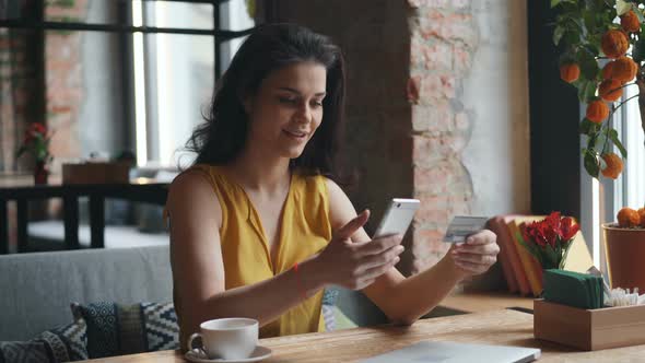 Young Woman Making Online Payment with Smartphone in Cafe Holding Bank Card