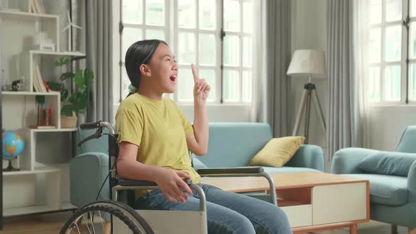Asian Kid Girl Sitting In A Wheelchair And Thinking Then Raising Her Index Finger In Living Room