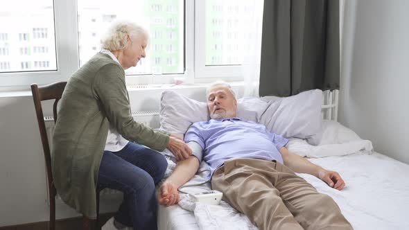 an Elderly Woman Checks Her Husband's Blood Pressure with a Blood Pressure Monitor