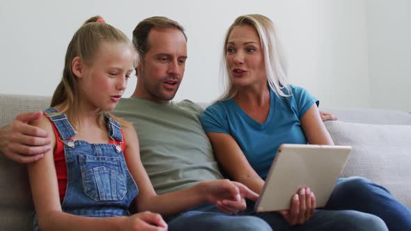 Caucasian parents sitting on sofa with daughter talking and looking at digital tablet