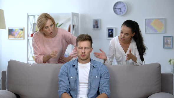 Tired Man Sitting on Sofa, Enduring to Wife and Mother Criticize Him, Quarrel