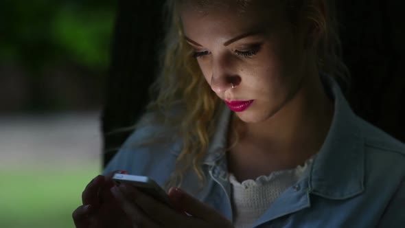 Young woman tapping screen of smart phone hand hold, face illuminated by screenlight.