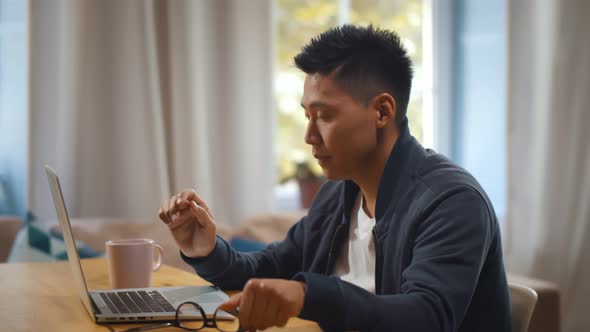 Tired Asian Man Putting Off Glasses and Rubbing Eyes Working on Laptop in Home Office