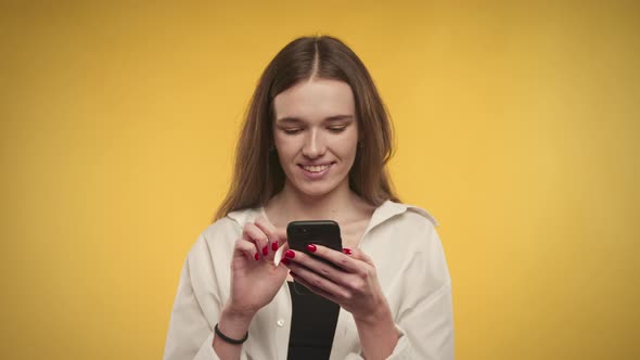 Young Adult Caucasian Woman is Typing on Her Smartphone on a Bright Yellow Background